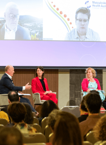 The image shows several members of a panel discussion on stage. Two people are joining the conversation via video call and can be seen on screen above. 